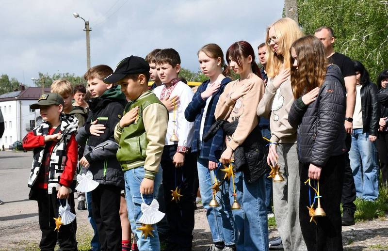 Група дітей та молоді стоїть на вулиці під час заходу пам'яті, притиснувши праву руку до серця під час виконання гімну. В руках вони тримають символічні паперові янголята, золоті зірочки та дзвоники.
