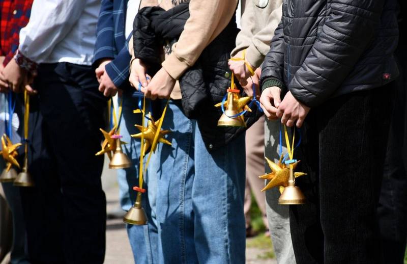 Крупний план рук дітей та підлітків, які тримають паперових білих янголят, золотисті декоративні зірочки та маленькі дзвоники на синьо-жовтих стрічках під час комеморативного заходу.