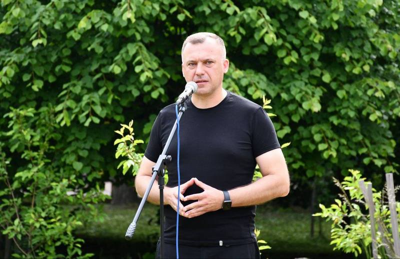 Макарівський селищний голова Вадим Токар виступає з промовою біля мікрофона на тлі зелених дерев під час заходу, присвяченого вшануванню пам'яті загиблих дітей.