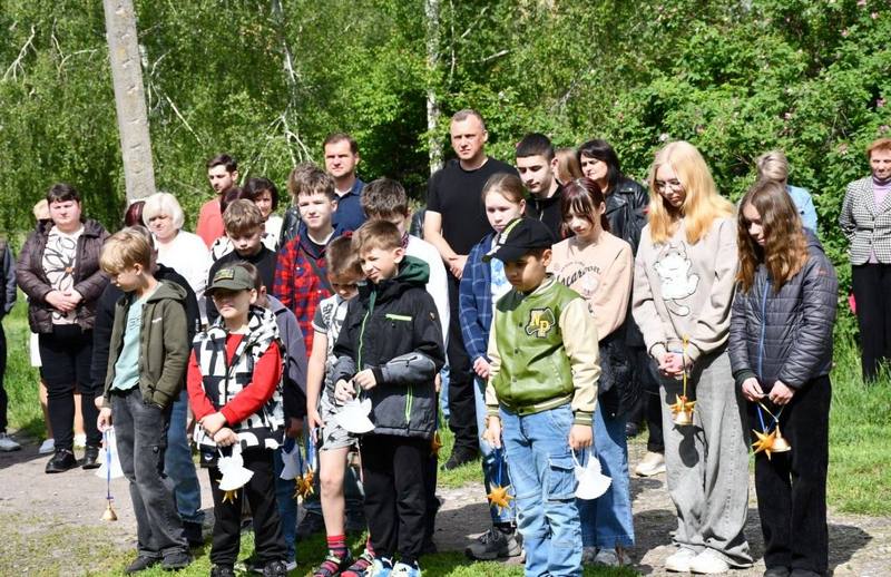 Жителі громади, дорослі та діти, стоять на подвір'ї під час мітингу-реквієму до Дня вшанування пам'яті дітей, які загинули внаслідок війни. Багато дітей тримають у руках символічні дзвоники та янголят.
