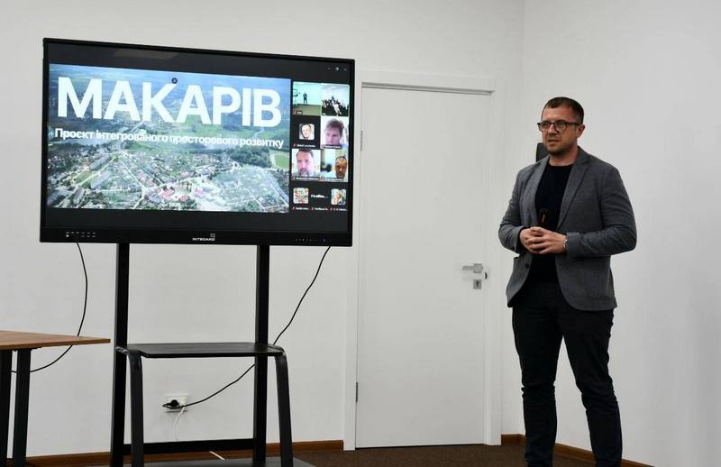 Анатолій Карбовський виступає з промовою перед аудиторією, стоячи поруч із великим презентаційним екраном з написом «МАКАРІВ. Проєкт інтегрованого просторового розвитку»