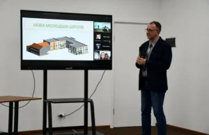 Спікер заходу коментує презентацію на великому екрані, де продемонстровано архітектурну тривимірну модель будівлі з написом «НОВА МОЛОДША ШКОЛА»