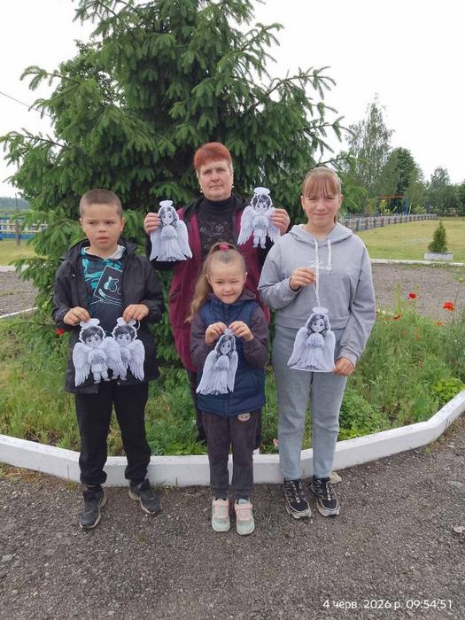 Троє учнів та вчителька стоять перед великою ялиною на вулиці, тримаючи паперових янголів для акції пам'яті «Голоси дітей»