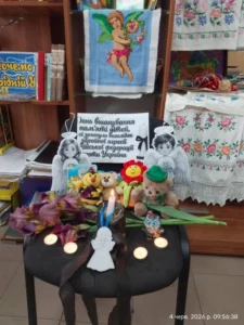 Крупний план меморіальної композиції на стільці: запалена свічка, м'які іграшки, квіти іриси та плакат про вшанування пам'яті дітей, загиблих внаслідок збройної агресії РФ