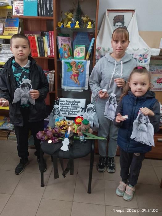 Троє дітей різного віку стоять у приміщенні бібліотеки, тримаючи в руках вирізані паперові зображення янголів. Перед ними на стільці облаштовано меморіал зі свічкою, квітами та іграшками