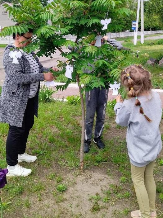 Вчителька та двоє дітей розвішують білих паперових янголів на гілках дерева на подвір'ї, вшановуючи пам'ять загиблих дітей України