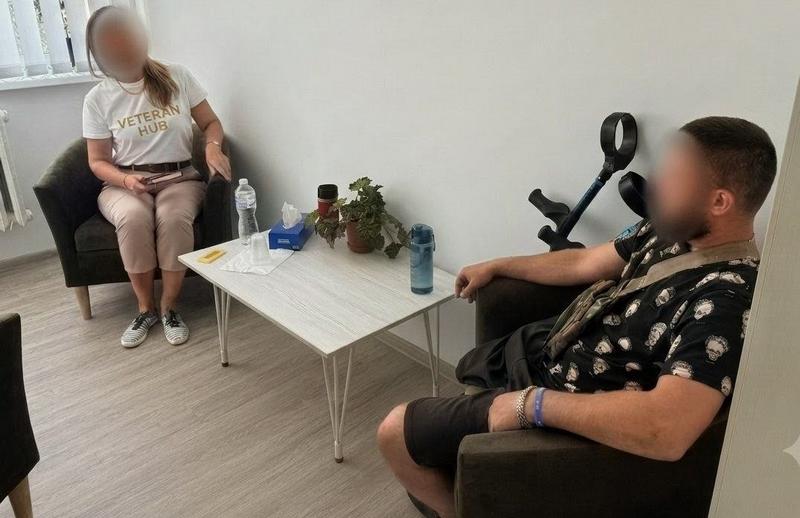 Психологічна підтримка військового. Захисник сидить у кріслі поруч із милицями та спілкується з фахівчинею у білій футболці з написом «Veteran Hub»