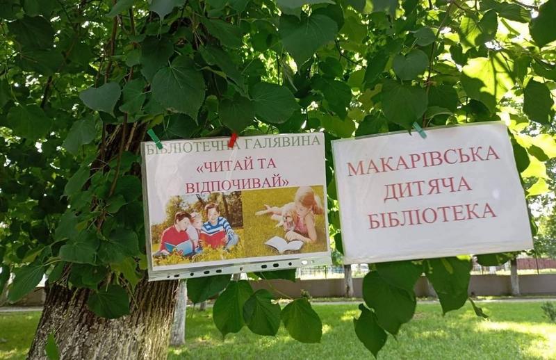 Два інформаційні плакати, закріплені прищіпками на гілках зеленого дерева. На плакатах написи: «Бібліотечна галявина "Читай та відпочивай"» з фотографіями дітей, що читають, та «Макарівська дитяча бібліотека»