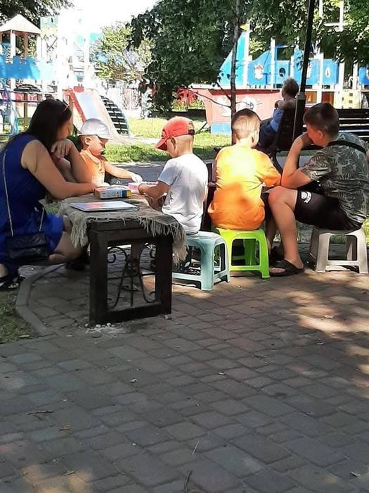 Група дітей та жінка сидять на маленьких кольорових стільчиках навколо столика на брукованій алеї парку, займаючись настільними іграми чи творчістю на свіжому повітрі. На фоні видно дитячий майданчик