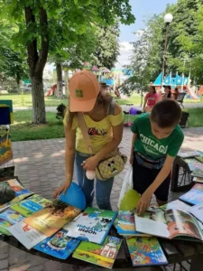 Дівчина в кепці та хлопчик у зеленій футболці розглядають розгорнуті дитячі книжки, що розкладені на лавці у літньому парку. Серед книг видно обкладинку коміксу про Смурфів
