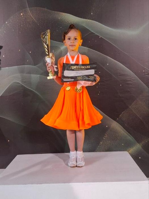 Юна чемпіонка Ясміна Мельник у яскраво-помаранчевій сукні стоїть на білому п'єдесталі пошани. На шиї дівчинки блищать золоті медалі, у руках вона гордо тримає свій заслужений кубок та диплом переможця Всеукраїнських змагань.