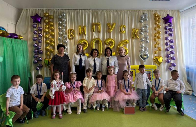 Велика спільна фотографія випускників Кодрянського ліцею разом із вихователями та педагогами у святковій залі. Діти у вишиванках та вечірніх вбраннях сидять і стоять у ряд, а позаду них усміхаються дорослі на фоні білої завіси, прикрашеної гірляндами з різнокольорових кульок та великим золотим написом «ВИПУСК 2026».