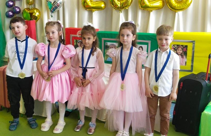 Юні випускники груп «Веселка» та «Сонечко» стоять у ряд під час урочистого свята. Дівчатка вдягнені у пишні святкові сукні рожевих та пастельних відтінків з білими бантами у волоссі, хлопчики — у світлі сорочки. На грудях у кожного вихованця гордо блищить золота медаль випускника дитячого садка на синій стрічці.