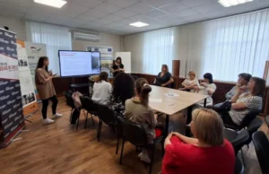 Робочий момент тренінгу з протидії насильству. Дві фахівчині стоять біля великого інтерактивного екрана з презентацією та виступають перед аудиторією. Учасники навчання сидять навколо великого столу і слухають доповідачок.