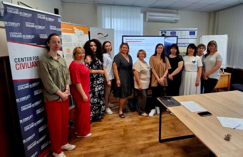 Групове фото учасниць та організаторок навчання у Макарівській громаді. Жінки стоять в один ряд у робочому кабінеті на тлі презентаційного екрана та інформаційних банерів із написами «Center for Civilians in Conflict» (CIVIC)