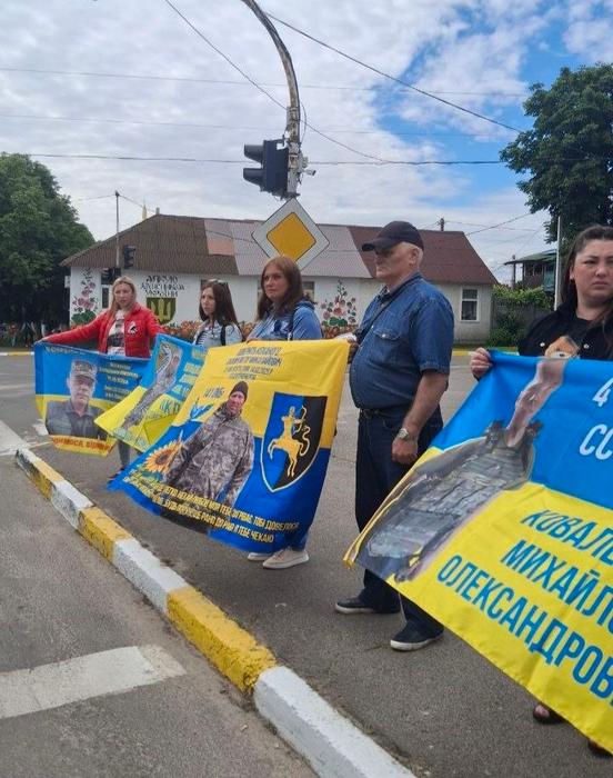 Учасники мирної акції стоять уздовж дороги та тримають великі синьо-жовті банери з портретами, іменами та військовою символікою зниклих безвісти захисників України
