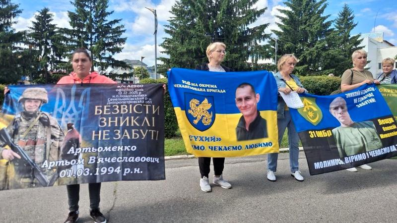 Рідні військових тримають великі плакати: ліворуч — банер пам'яті Артьоменка Артьома з написом «Безвісти зниклі не забуті», по центру — прапор 158-ї бригади з портретом воїна на псевдо «Сухий».