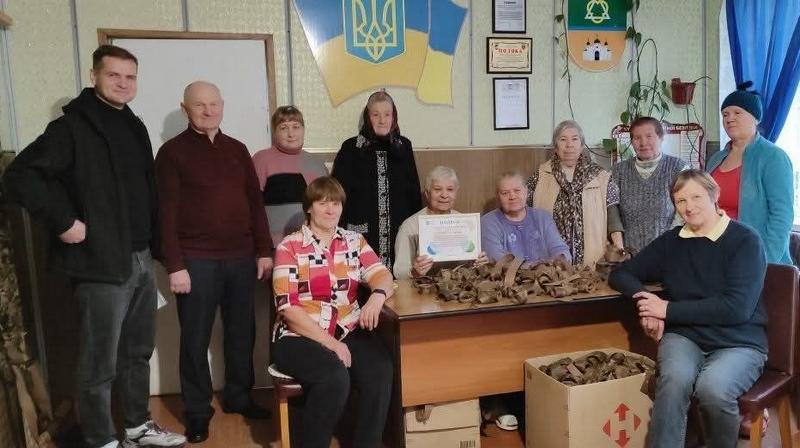 Команда волонтерів старостату (переважно літні люди) зібралася навколо робочого столу, одна з учасниць майстерні тримає в руках спільну грамоту-подяку за допомогу армії.