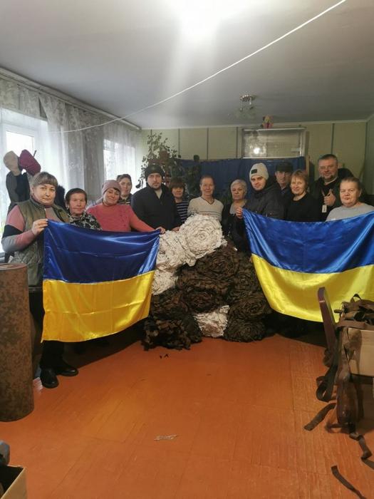 Велика група волонтерів майстерні села Великий Карашин позирує разом у кімнаті, тримаючи в руках два розгорнуті державні прапори України на тлі великої кількості готових маскувальних сіток.