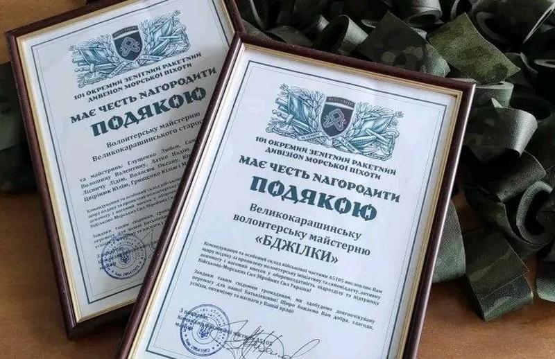Дві офіційні подяки у дерев'яних рамках на тлі камуфляжних стрічок, вручені волонтерській майстерні «Бджілки» та майстриням Великокарашинського старостату від 101 окремого зенітного ракетного дивізіону морської піхоти.