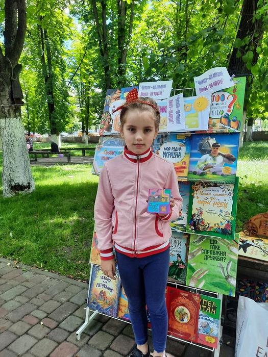 Маленька дівчинка в рожевій кофті тримає в руках саморобку та позує на тлі яскравого стенда з дитячими книжками у парку.