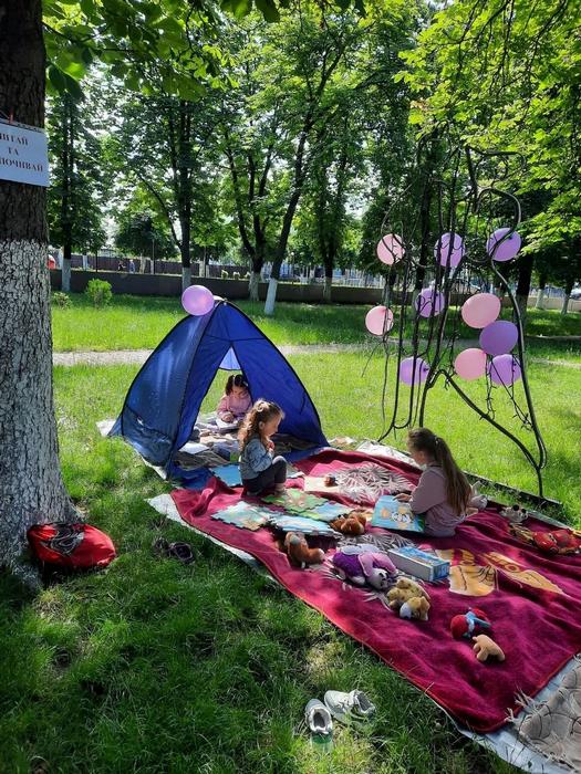 Дитяча зона відпочинку у зеленому парку. Дівчатка граються та читають книги у синьому ігровому наметі та на великому м'якому пледі, розстеленому на траві.