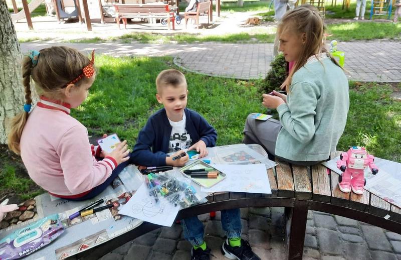 Троє дітей сидять на дерев'яній лавці у сквері та захоплено малюють фломастерами на свіжому повітрі