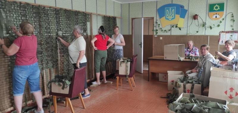 Процес роботи у волонтерській майстерні: декілька жінок стоять уздовж стіни та вплітають захисні стрічки у велику маскувальну сітку, поруч стоять коробки з матеріалами.