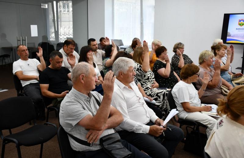 Члени виконавчого комітету Макарівської селищної ради сидить на стільцях у залі засідань та піднімають руки в процесі голосування за важливі питання громади.