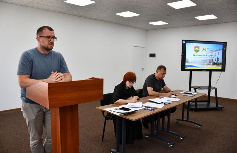 Карбовський А.Г. виступає за дерев'яною трибуною під час засідання виконкому. На задньому плані за столом сидять секретар, яка робить записи, та Макарівський селищний голова Вадим Токар.
