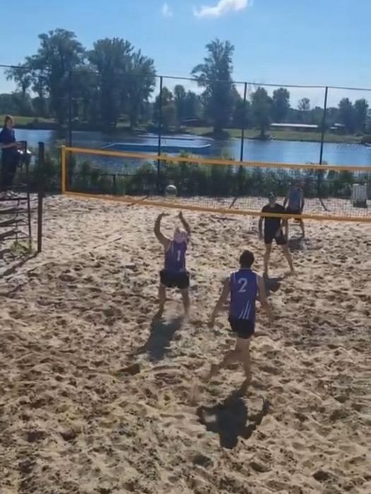 Двоє гравців у фіолетових майках (один під номером "1", інший під номером "2") перебувають на піщаному майданчику для пляжного волейболу. Позаду, на суддівській вишці, сидить людина, а на задньому плані видніється річка. Ще два гравці знаходяться на протилежному боці сітки під час гри на турнірі “Beginner’s” у Києві.