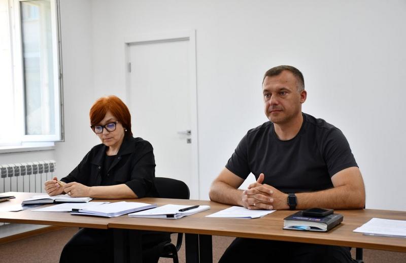 Макарівський селищний голова Вадим Токар сидить за робочим столом поруч із секретарем виконкому. На столі перед ними лежать документи, нотатники та ручки.