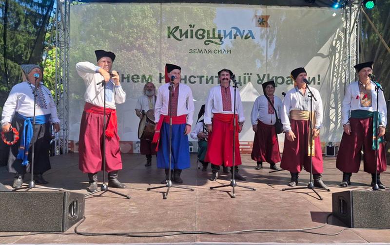 Народний аматорський фольклорний колектив «Козацькі джерела» у шароварах, вишиванках та кучмах виступає на головній сцені фестивалю на тлі банера «Київщина — земля сили».