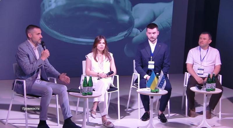 Чотири спікери — три чоловіки та одна жінка — під час панельної дискусії на презентації антикорупційного гайду. Чоловік ліворуч говорить у мікрофон. На столиках стоять пляшки з водою та маленькі прапори України й ОБСЄ