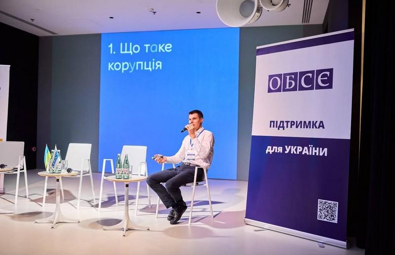 Спікер у білій сорочці сидить на стільці на сцені та виступає з мікрофоном на презентації гайду. На великому синьому екрані позаду нього відображено слайд із заголовком «1. Що таке корупція». Праворуч стоїть банер ОБСЄ з написом «ПІДТРИМКА ДЛЯ УКРАЇНИ»