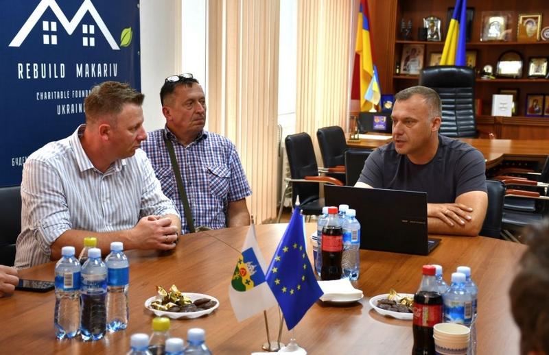 Офіційна зустріч у кабінеті: Макарівський селищний голова виступає перед польськими колегами за круглим столом із прапорами України, Польщі та ЄС. На стіні — банер благодійного фонду «Rebuild Makariv».