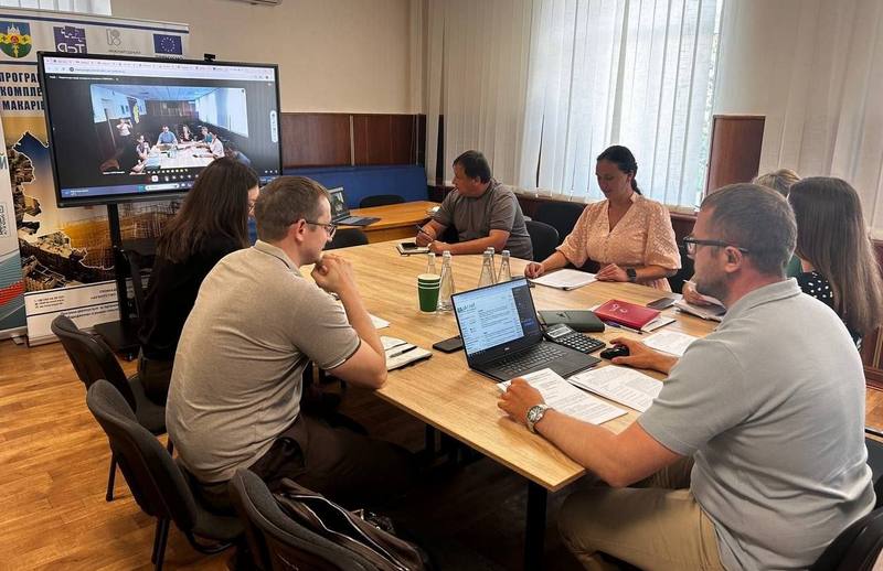Робоча зустріч у Макарівській селищній раді щодо проєкту HOPE. Учасники обговорюють документацію за столом, на стіні — великий екран із відеозв'язком.