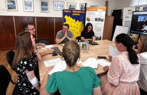 Процес переговорів у кабінеті селищної ради. Учасники зустрічі вивчають роздруковані документи. На стіні позаду видно інформаційні стенди та карту Макарівського району в жовто-синіх кольорах.