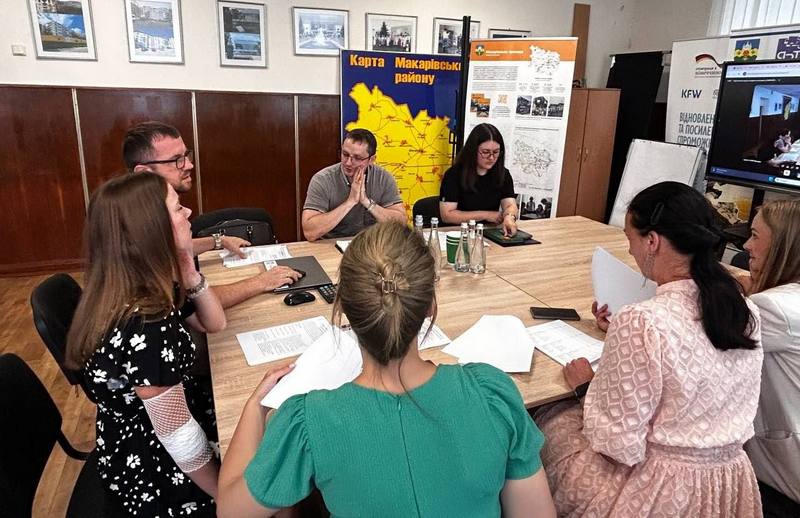 Процес переговорів у кабінеті селищної ради. Учасники зустрічі вивчають роздруковані документи. На стіні позаду видно інформаційні стенди та карту Макарівського району в жовто-синіх кольорах.