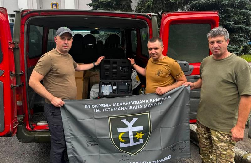 Юрій Сірцов разом із волонтерами та військовими стоять біля відчиненого багажника червоного мікроавтобуса, де лежить відкритий кейс із квадрокоптером Autel EVO Max 4T. Вони тримають прапор 54-ї окремої механізованої бригади імені гетьмана Івана Мазепи з підписами.