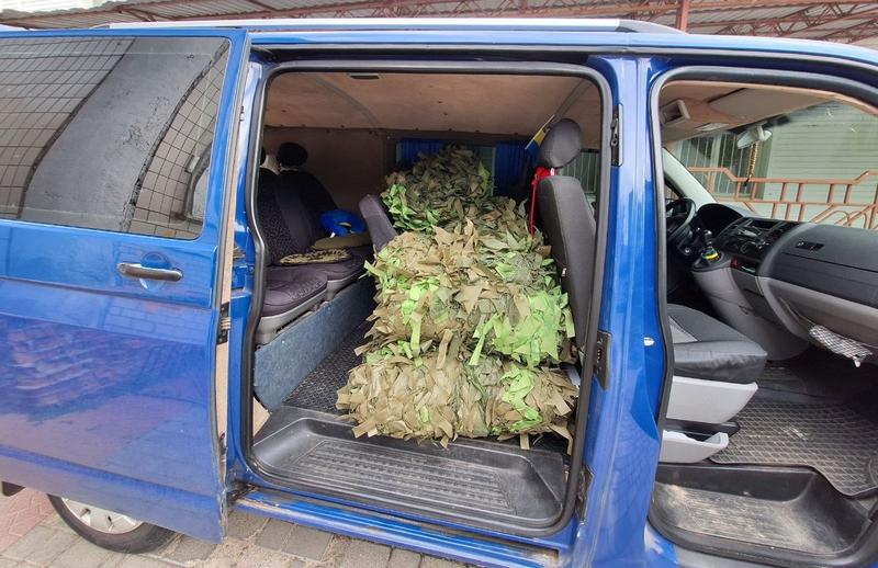 Салон синього мікроавтобуса з відкритими боковими дверима, повністю завантажений сплетеними зеленими та коричневими маскувальними сітками для українських захисників.