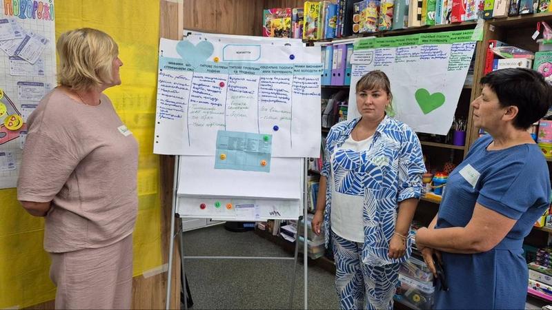 Три учасниці групової супервізії обговорюють робочі завдання біля фліпчарту з таблицею потреб сімей