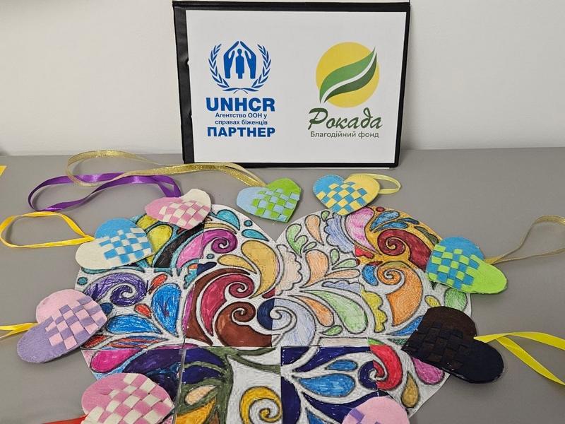 Творчі вироби учасників сімейної зустрічі: велике розфарбоване серце-пазл та маленькі плетені серця з фетру на тлі логотипів БФ «Рокада» та UNHCR.