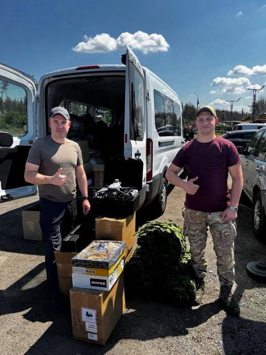 Заступник селищного голови Юрій Сірцов разом із волонтером показують великий палець вгору біля буса з розвантаженими дронами та коробками.