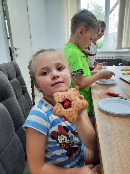 Дівчинка з косичками, одягнена в синьо-білу смугасту футболку з малюнком Міккі Мауса, сидить за столом і дивиться в камеру. Вона тримає в руці розписаний пряник у формі зірочки. Поруч за столом видно інших дітей, які продовжують роботу.