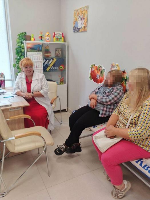 Лікарка у білому халаті за робочим столом веде дружню розмову з дитиною та її матір'ю, які сидять навпроти.
