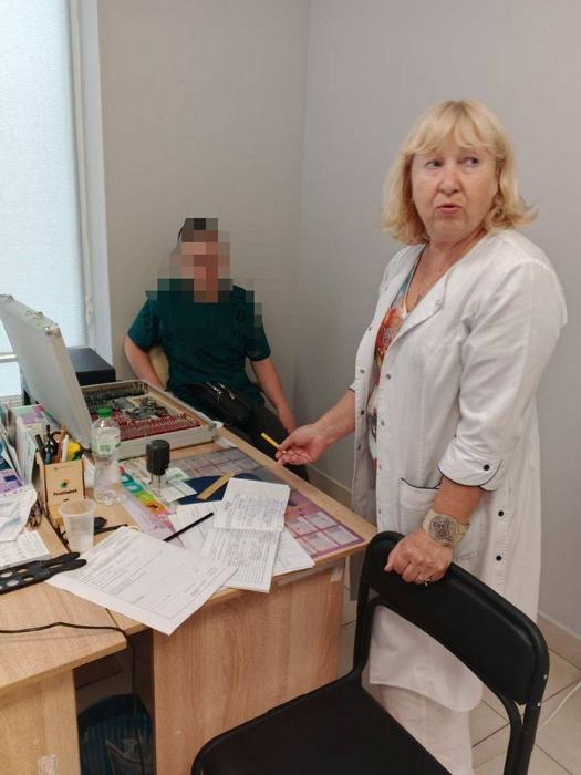Лікарка-офтальмолог у білому халаті стоїть біля столу з обладнанням для перевірки зору, консультуючи підлітка.