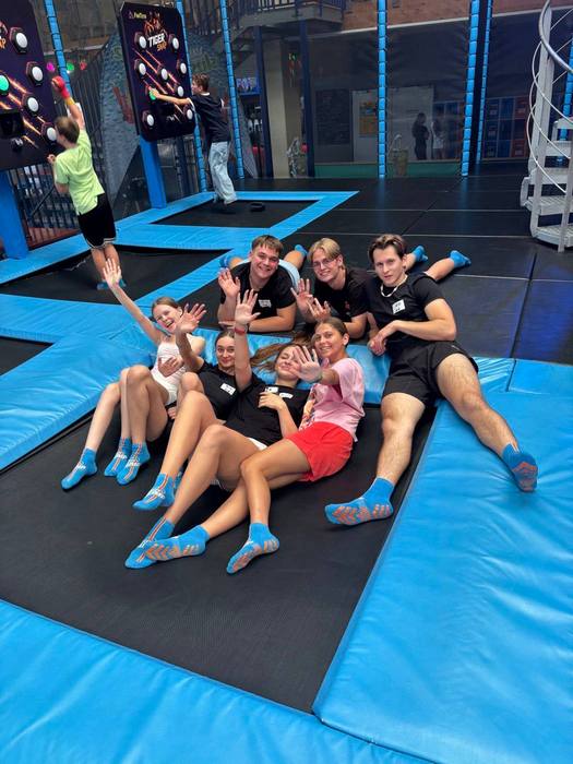 Група молоді у синіх шкарпетках лежить на синьому батуті в Jump Park та махає руками в камеру.