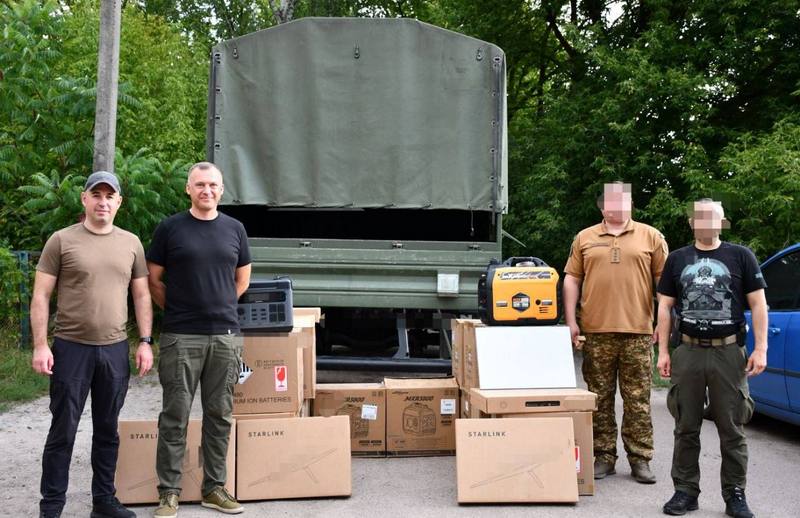 Представники Макарівської громади та військові (із замаскованими обличчями) стоять перед вантажівкою. Перед ними розкладено коробки з написом 'Starlink', коробки з генераторами, один відкритий жовтий інверторний генератор та портативна зарядна станція.