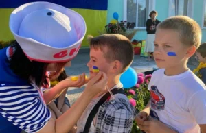 Знімок крупним планом, на якому дівчина-аніматор (у білому капелюсі з червоним написом та тільняшці) обережно наносить пензликом патріотичний аквагрим у кольорах українського прапора на щоку маленькому хлопчику в картатій сорочці. Поруч стоїть інший хлопчик із синім мазком на щоці.